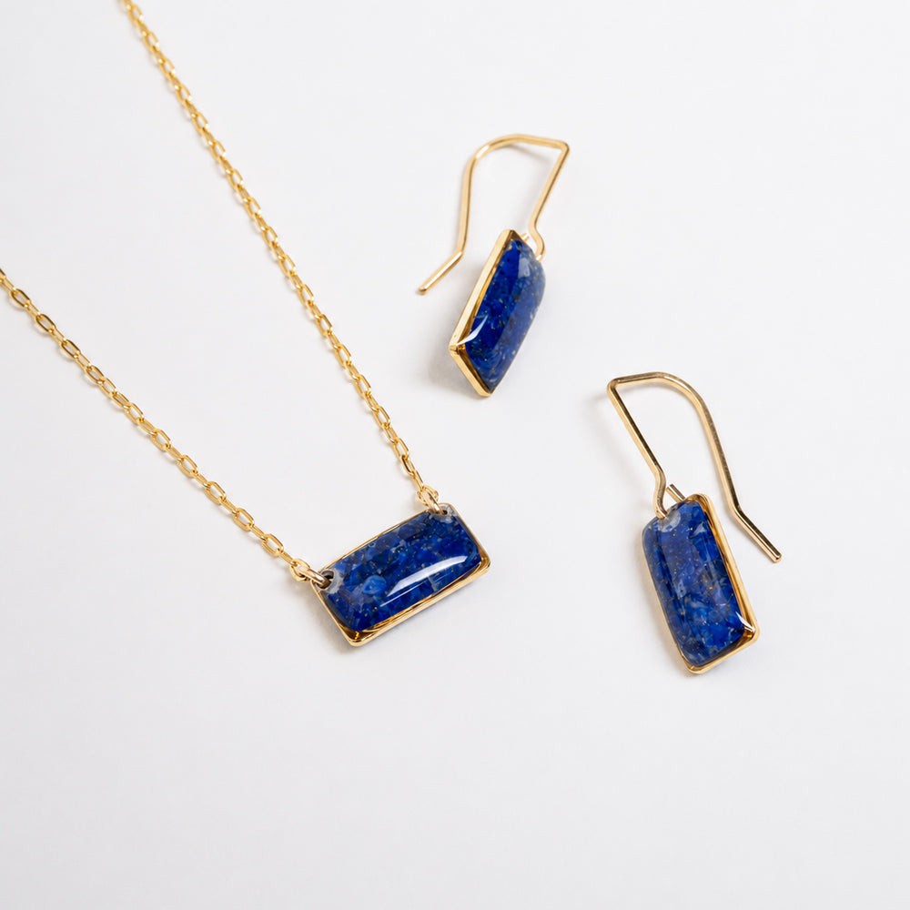 A delicate gold chain necklace featuring a small, rectangular blue stone pendant with gold bezel settings, shown against a clean off-white background.