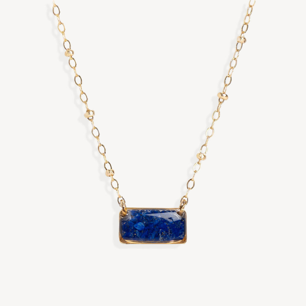 A delicate gold chain necklace featuring a small, rectangular blue stone pendant with gold bezel settings, shown against a clean off-white background.