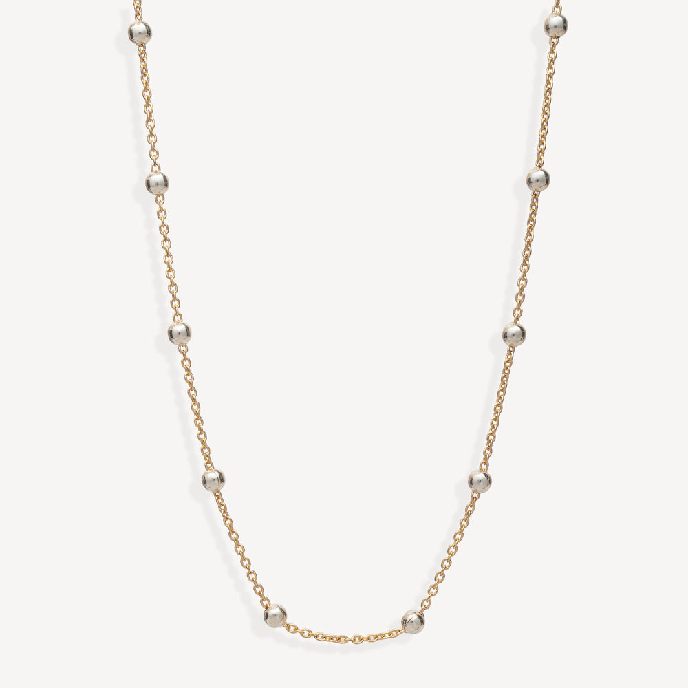A delicate gold chain necklace featuring evenly spaced, polished silver-toned round beads, displayed against a clean off-white background.