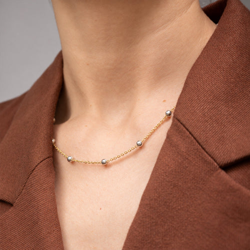 A delicate gold chain necklace with small silver-toned bead accents, shown worn on a model's neck over a brown blazer.