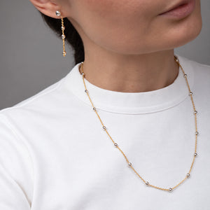 A close-up of a model wearing a delicate gold beaded necklace and a matching drop earring, paired with a white crewneck top against a neutral gray background.