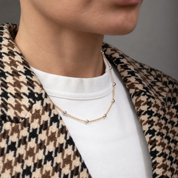 A close-up of a person wearing a delicate gold chain necklace with small silver bead accents, styled over a white t-shirt and a brown, beige, and black houndstooth blazer.
