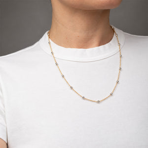 A delicate gold chain necklace with small silver bead accents, shown worn on a model's neck over a white t-shirt.