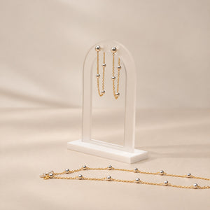 Gold beaded jewelry set displayed on a clear acrylic stand, featuring matching drop earrings and a long necklace, photographed on a warm beige background.
