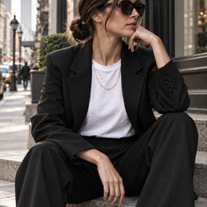 A woman sits on outdoor stone steps wearing a tailored black blazer and matching wide-leg pants over a white crew-neck T-shirt, with gold jewelry and dark sunglasses. The background shows a softly blurred city street scene.