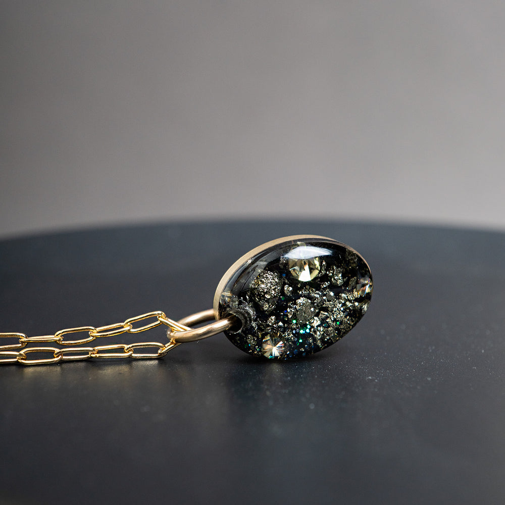 Gold chain necklace with an oval pendant containing black mineral inclusions and a small iridescent stone, shown on a light gray background.
