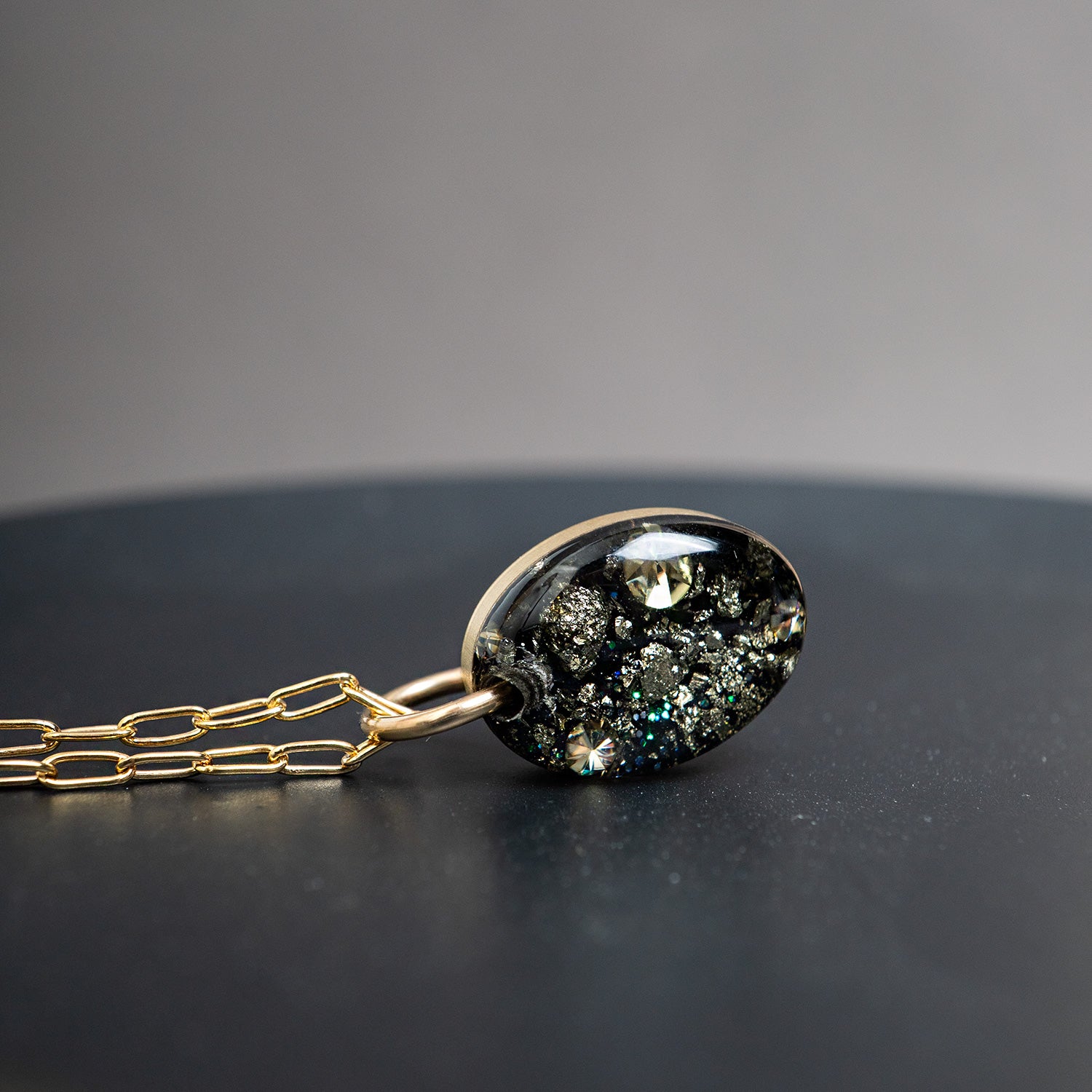 A gold chain necklace with an oval pendant featuring a black background filled with sparkling gold, silver, and iridescent flecks, resting on a dark surface against a gray background.
