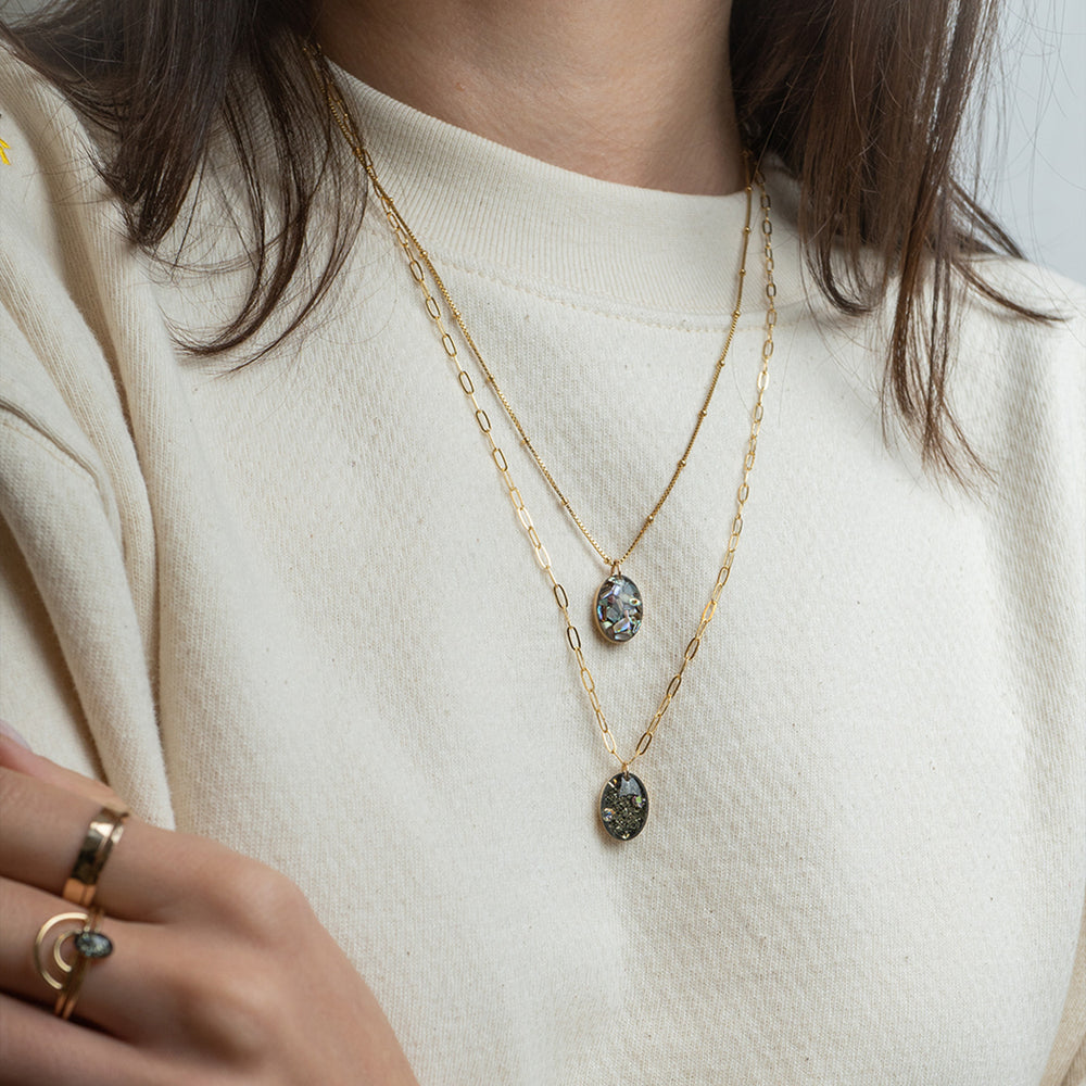 Gold chain necklace with an oval pendant containing black mineral inclusions and a small iridescent stone, shown on a light gray background.