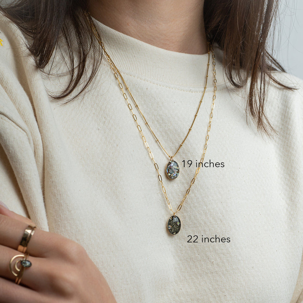 Gold chain necklace with an oval pendant containing black mineral inclusions and a small iridescent stone, shown on a light gray background.