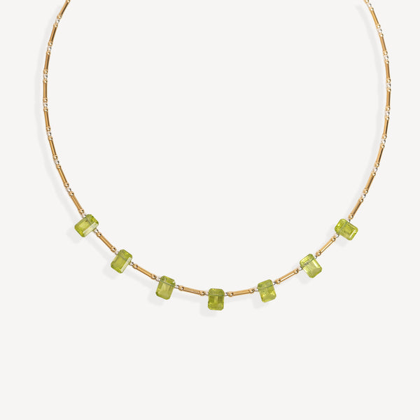 A delicate gold chain necklace featuring a row of small, faceted green gemstones spaced along the front, displayed against a clean, light off-white background.