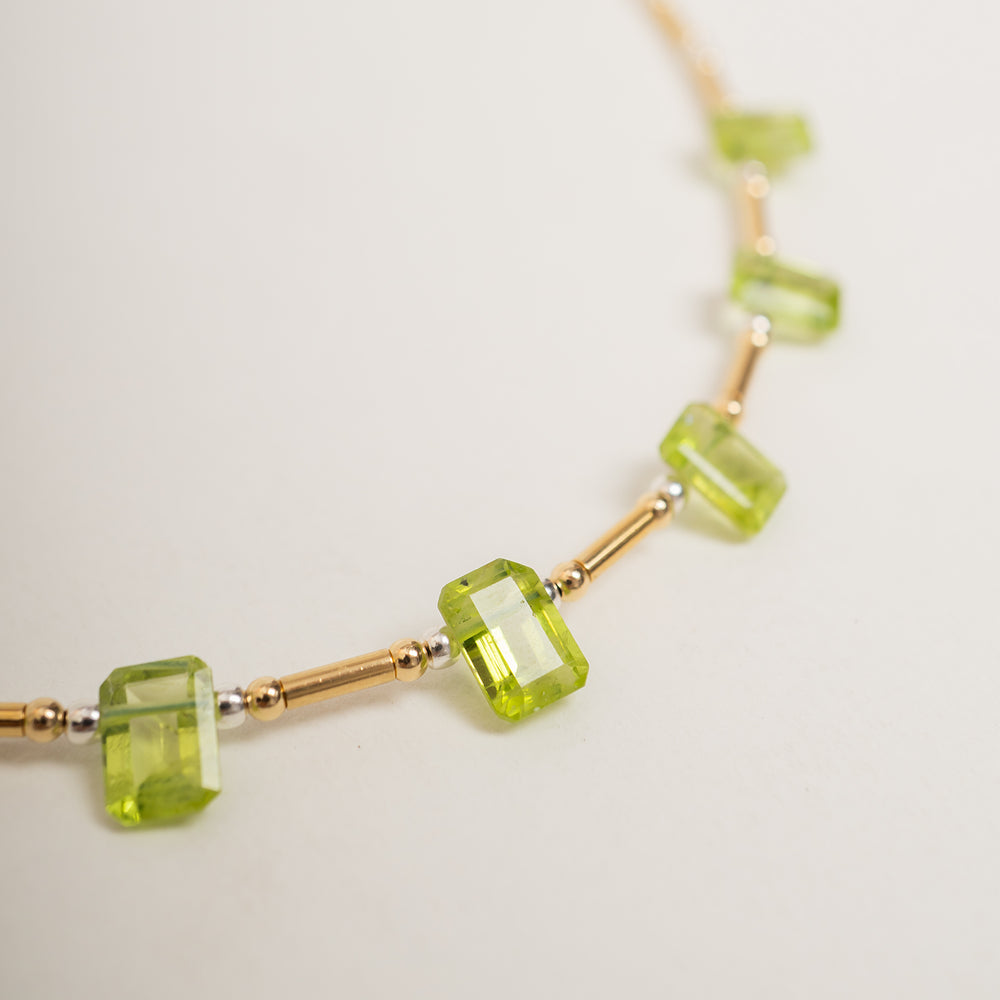 A delicate gold chain necklace featuring a row of small, faceted lime green gemstone beads, displayed against a clean off-white background.