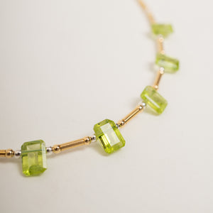 A close-up of a necklace featuring faceted light green gemstone beads set on a gold-toned chain with small silver spacer beads, photographed on a plain light background.