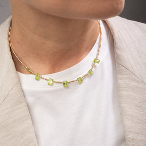 A close-up of a person wearing a delicate gold chain necklace with small, rectangular light green gemstones, styled over a white t-shirt and a light beige blazer.