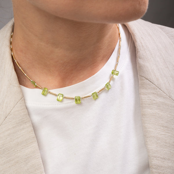A close-up of a person wearing a delicate gold chain necklace with small, rectangular light green gemstones, styled over a white t-shirt and a light beige blazer.