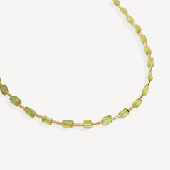 A delicate gold chain necklace featuring small, faceted light green gemstone beads, photographed against a clean off-white background.
