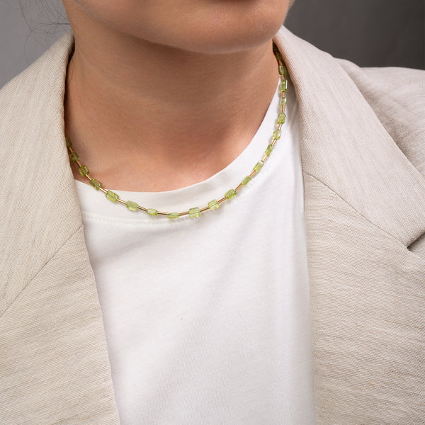 A close-up of a person wearing a delicate beaded necklace made of small light green faceted beads, styled over a white t-shirt and a light beige blazer.