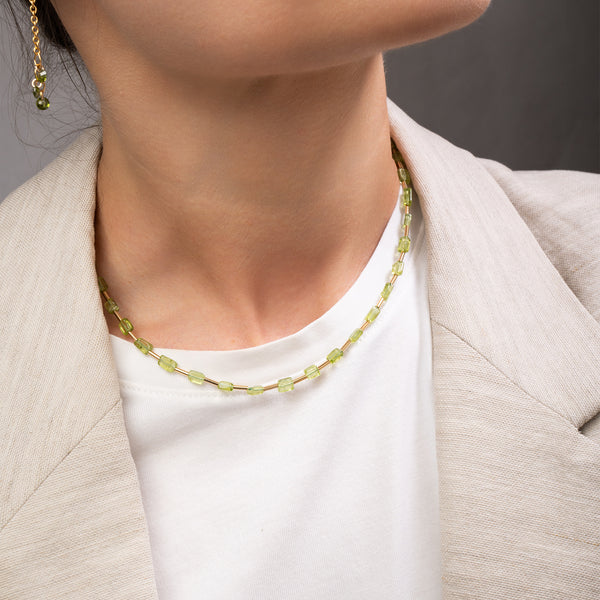 A close-up of a model wearing a delicate gold chain necklace with small, light green faceted beads, styled with a white t-shirt and a beige blazer.