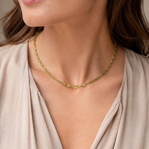 A close-up of a woman wearing a delicate gold chain necklace with small green gemstone accents, paired with a beige blouse.