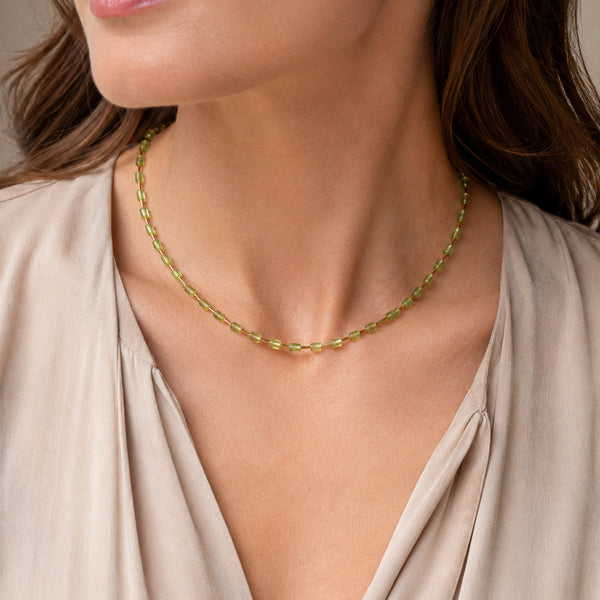 A close-up of a woman wearing a delicate gold chain necklace with small green gemstone accents, paired with a beige blouse.