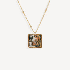 A gold chain necklace with a square pendant featuring a detailed, colorful floral and nature illustration, set against a plain off-white background.
