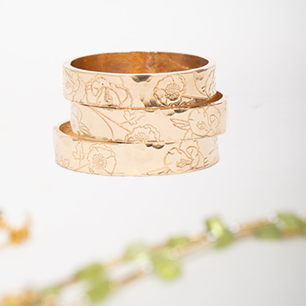 A gold band ring with a hammered texture and engraved floral patterns, shown on a light gray background.