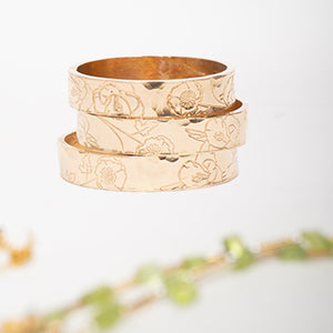 Three stacked gold-toned rings with an engraved floral pattern, displayed on a clean white background with a soft-focus decorative plant in the foreground.