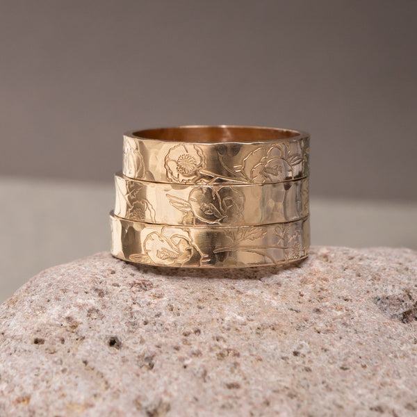 A stack of three wide gold-toned rings with embossed floral patterns, displayed on a textured stone surface against a neutral gray background.