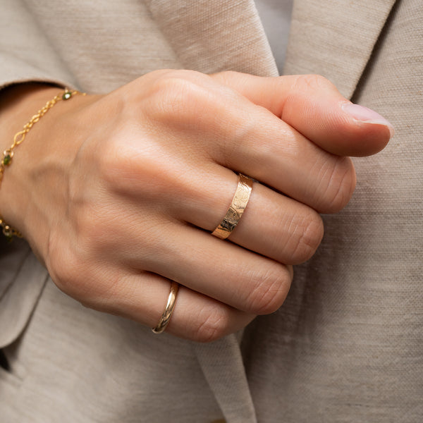 A close-up of a person's hand wearing two gold rings, one textured band ring and one slim twisted ring, with a gold bracelet on the wrist, set against a beige blazer.
