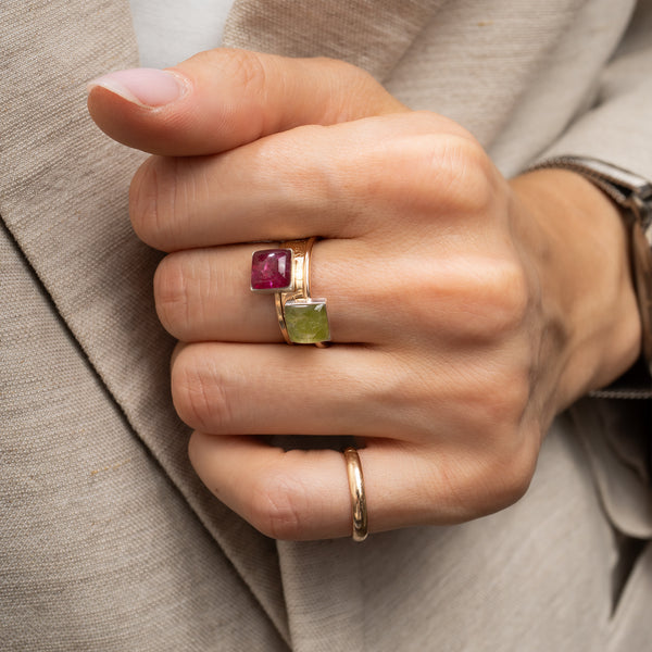 A close-up of a hand wearing a gold ring set with a square pinkish-red gemstone and a square green gemstone, with a simple gold band on another finger, against a beige blazer background.