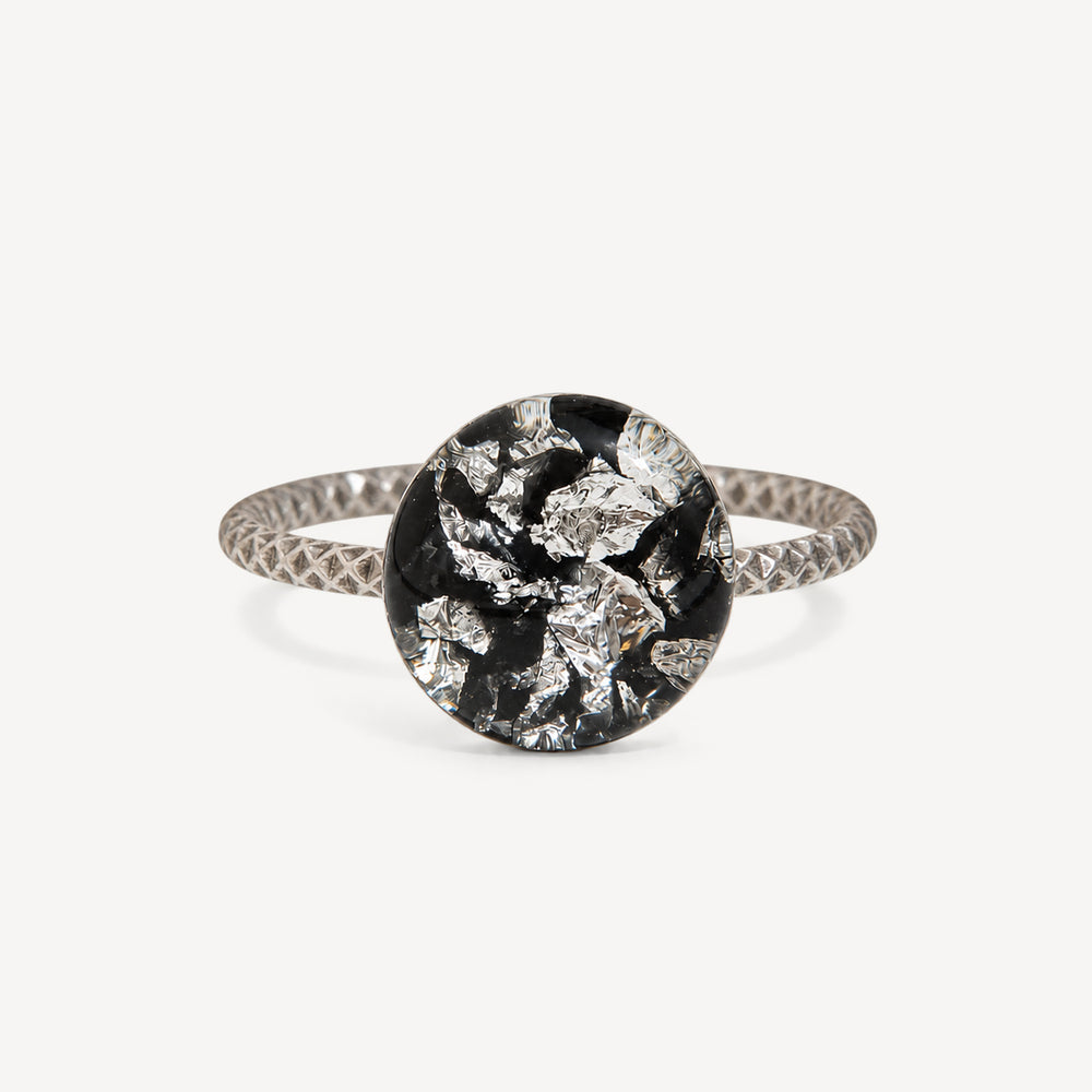 A silver ring with a textured band and a round bezel-set black and silver foil mosaic center, shown on a light gray background.