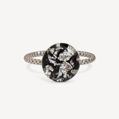 A silver ring with a textured band and a round bezel-set black and silver foil mosaic center, shown on a light gray background.