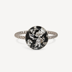 A silver ring with a textured band and a round bezel-set black and silver foil mosaic center, shown on a light gray background.