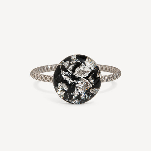A silver ring with a textured band and a round bezel-set black and silver foil mosaic center, shown on a light gray background.