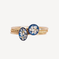 A gold ring stack featuring two oval blue resin cabochons with pressed white flowers, set against a plain light gray background.