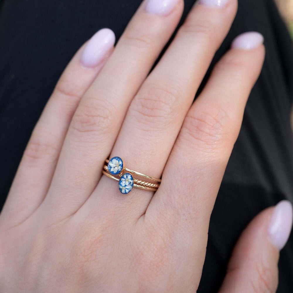 A gold ring stack featuring two oval blue resin cabochons with pressed white flowers, set against a plain light gray background.