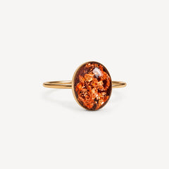 A gold ring with an oval cabochon featuring a textured, metallic copper-orange pattern, set against a clean off-white background.