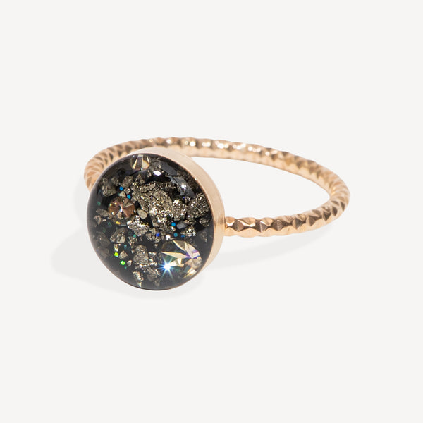 A gold ring with a textured band and a round bezel containing black resin, silver glitter, and iridescent crystals, set against a light gray background.