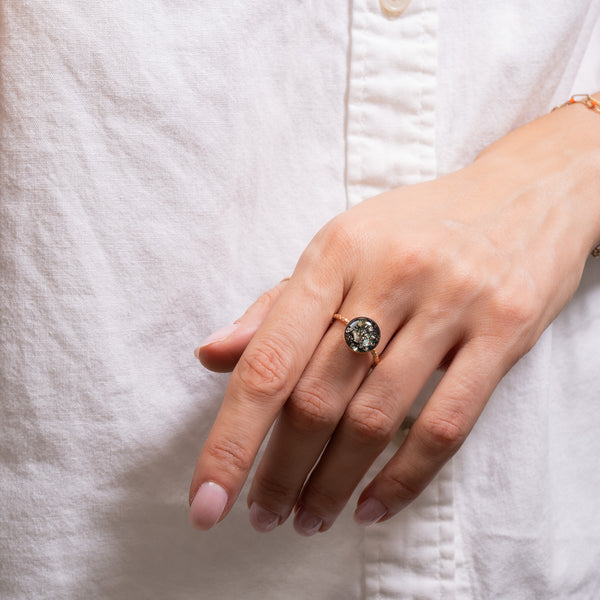 A gold ring with a textured band and a round bezel containing black resin, silver glitter, and iridescent crystals, set against a light gray background.