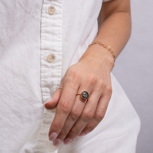 A close-up of a person's hand wearing a gold ring with a dark, textured oval centerpiece and a delicate gold chain bracelet, styled with a white button-up shirt.