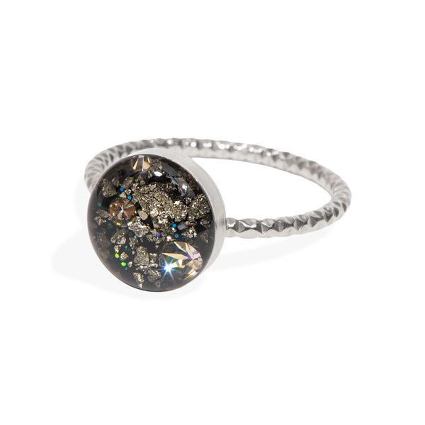 A silver ring with a textured band and a round bezel containing a black background filled with gold glitter, small crystals, and a prominent iridescent starburst rhinestone, shown on a white background.