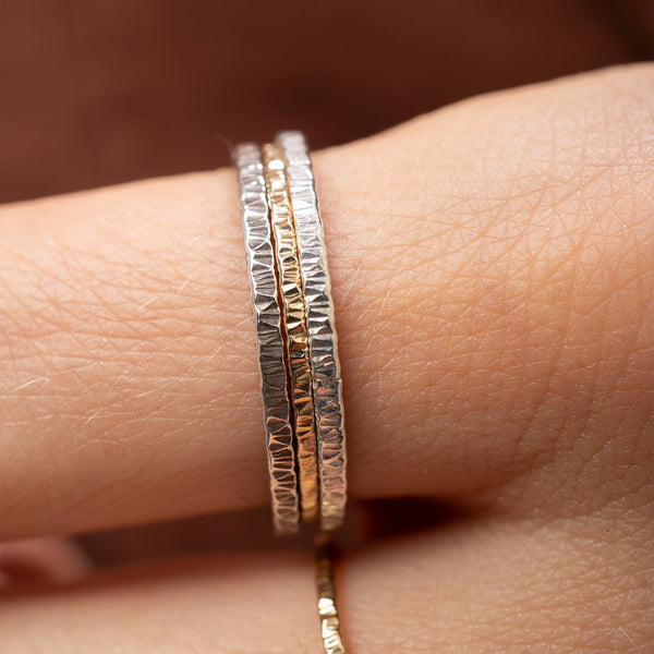 A close-up of a hand wearing three textured metal rings, including two silver rings and one gold ring, with a blurred warm brown background.