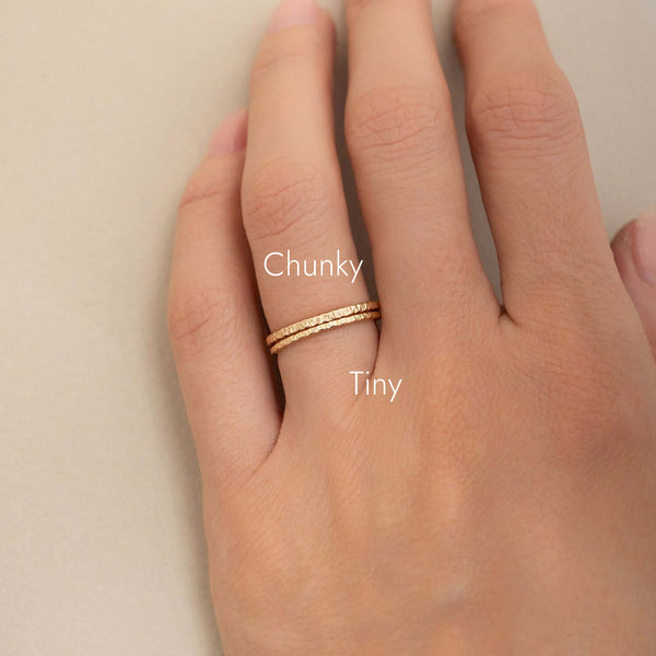 A close-up of a hand wearing two thin gold rings, with one chunkier textured ring labeled 'Chunky' and one thinner textured ring labeled 'Tiny'.