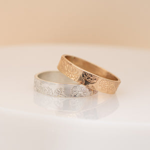 Two engraved rings, one in yellow gold and one in silver, resting on a reflective white surface against a soft beige background.
