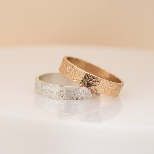 Two engraved rings, one in yellow gold and one in silver, resting on a reflective white surface against a soft beige background.