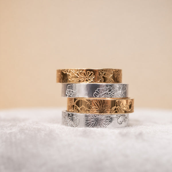 Four stacked rings with engraved floral patterns in gold and silver tones, resting on a soft white textured surface against a warm beige background.