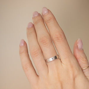 A close-up of a hand wearing a simple hammered silver band ring on the ring finger, with a second thin gold band ring on the back of the hand, set against a solid beige background.