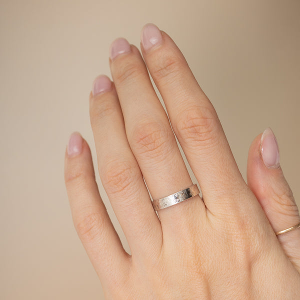 A close-up of a hand wearing a simple hammered silver band ring on the ring finger, with a second thin gold band ring on the back of the hand, set against a solid beige background.