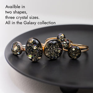 A set of gold-toned jewelry pieces with dark, glittery crystal-like droplets, displayed on a black plate against a light gray background. Text in the upper left reads: 'Available in two shapes, three crystal sizes.