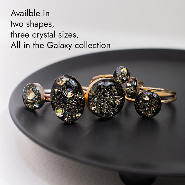A set of gold-toned jewelry pieces with dark, glittery crystal-like droplets, displayed on a black plate against a light gray background. Text in the upper left reads: 'Available in two shapes, three crystal sizes.