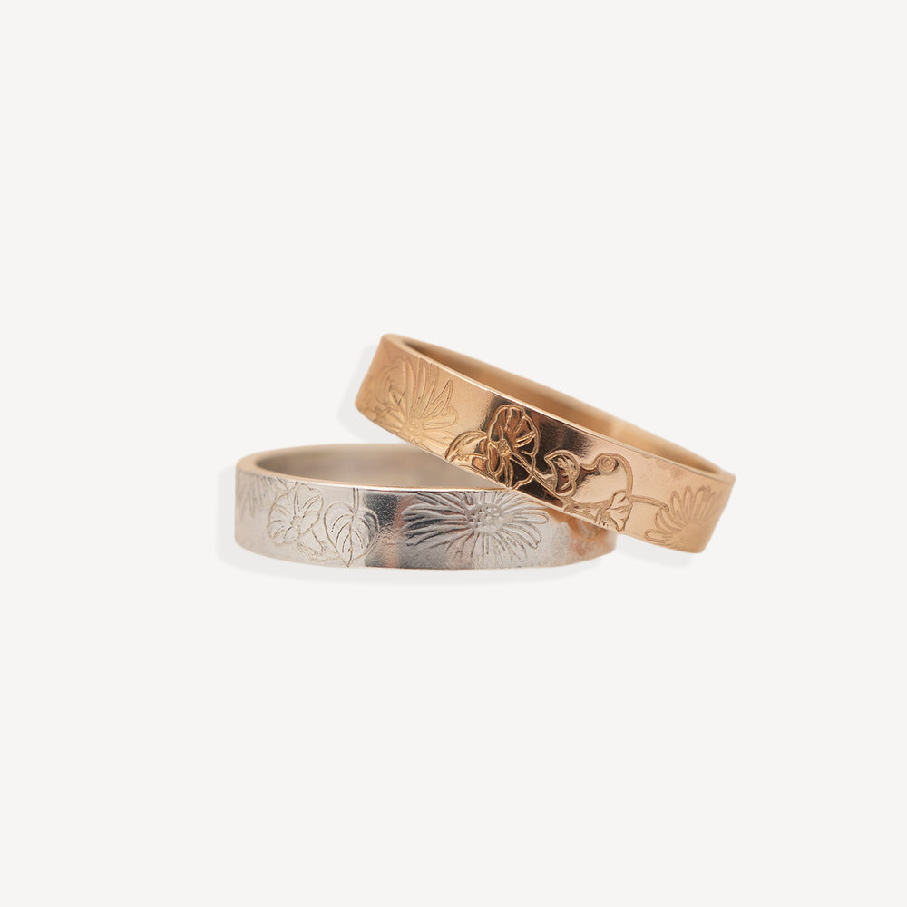Two engraved metal bands, one rose gold and one silver, overlapping on a clean off-white background. Both bands feature a floral pattern engraved on the outer surface.